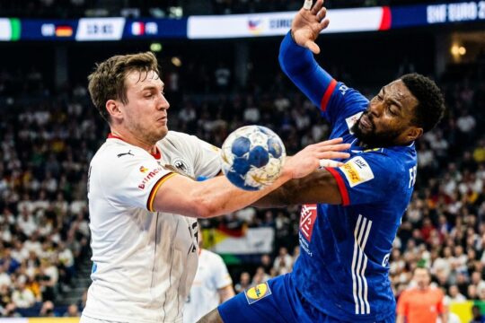 Ein deutscher Handballspieler wirft kraftvoll aufs Tor, während ein französischer Spieler athletisch zum Block springt. Intensive Szene der Handball EM Deutschland Frankreich.