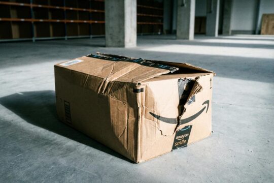 Stark zerknitterter, zerfetzter Amazon-Lieferkarton mit eingerissenem Smile-Logo auf kaltem Betonboden, symbolisiert den Amazon Stellenabbau.