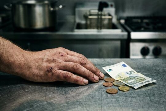 Erschöpfte Hand eines Gastronomiemitarbeiters auf Edelstahlarbeitsfläche, daneben nur wenige Cent. Mindestlohn Gastronomie Verstöße werden hier deutlich.
