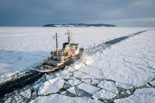 Ein mächtiger Hiddensee Eisbrecher durchbricht unter winterlichem Himmel dickes Ostsee-Eis, aufspritzende Schollen und eine dunkle Fahrrinne zur Insel Hiddensee.