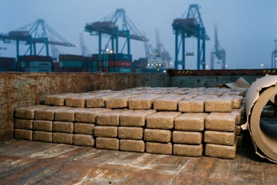 Dicht gepackte Heroin-Pakete auf rauem Metallgrund. Im unscharfen Hintergrund: Kräne und Container des Hamburger Hafens. Ein bedeutender Heroin Fund Hamburg und Bremen.