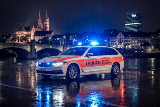 Ein Streifenwagen mit leuchtendem Blaulicht auf nassem Asphalt einer Basler Straße bei Nacht. Das Symbol für die Blaulicht Region Basel.