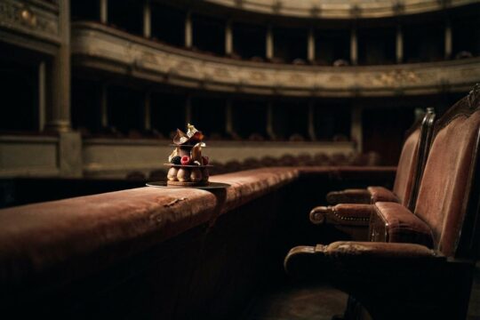 Ein kunstvolles Dessert balanciert prekär auf einem Theatersitz. Leere Reihen im Hintergrund zeigen die Kulturgastronomie Probleme und ihre fragile Existenz.