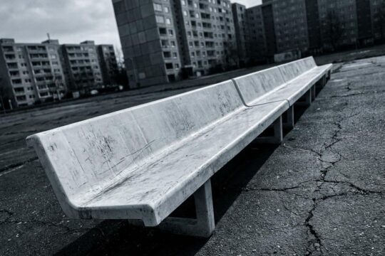 Abgenutzte weiße Plastikbank auf Asphalt vor Plattenbauten in Jena Lobeda. Die kühle Stimmung visualisiert das Theaterstück Hass Jena.