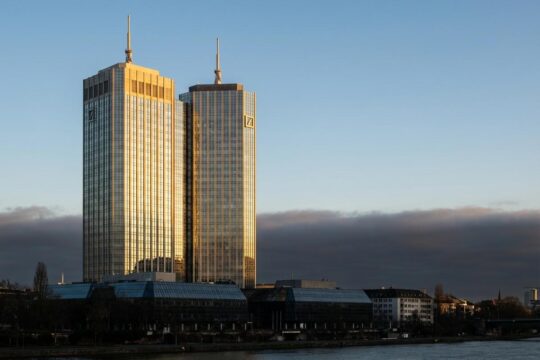 Die Zwillingstürme der Deutschen Bank in Frankfurt bei Sonnenaufgang, goldene Spitzen signalisieren den Rekordgewinn 2025. Schatten warnen vor Risiken.