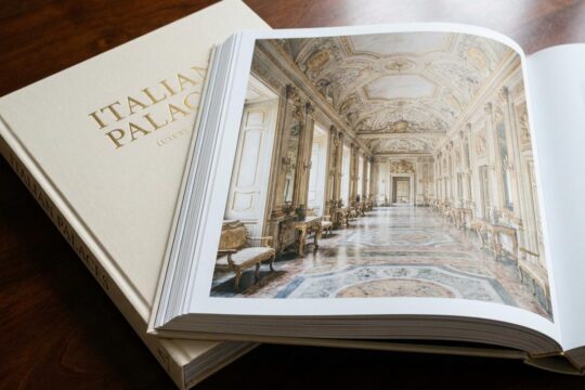 Ein aufgeschlagener Italian Palaces Bildband präsentiert eine majestätische Raumflucht eines italienischen Palastes mit Stuckdecken, goldenen Verzierungen und Marmorböden auf dunklem Holz.