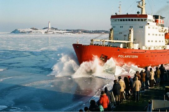 Ein mächtiger Eisbrecher nach Hiddensee pflügt durch die vereiste Ostsee. Passagiere stehen an Deck und blicken sehnsüchtig auf die schneebedeckte Insel.