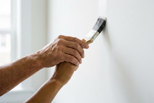 Zwei Hände unterschiedlicher Hauttöne streichen gemeinsam eine helle Wand. Ein Bild der harmonischen Zusammenarbeit und erfolgreichen Integration von Flüchtlingen in den Arbeitsmarkt.