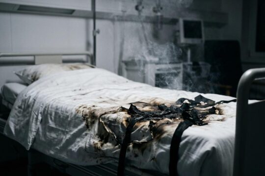 Nahaufnahme eines Krankenhausbettes mit verkohlter Bettwäsche und Brandspuren. Zerrissene Fixiergurte zeigen, wo ein Patient in Krankenhaus verbrennt. Leichter Rauch steigt auf.