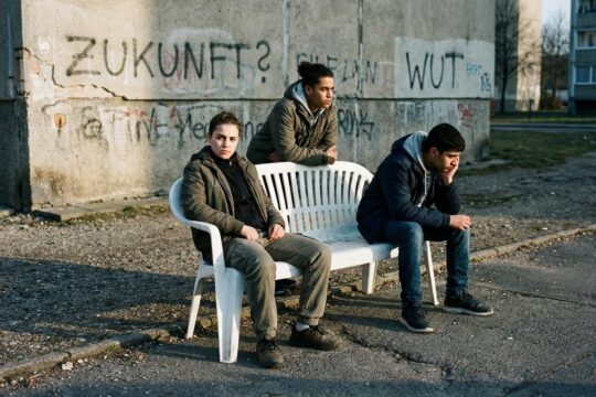 Drei junge Menschen auf ramponierter Bank vor Plattenbauwand, einer blickt herausfordernd. Spiegeln Entfremdung und Wut des Theaterhaus Jena Hass wider.