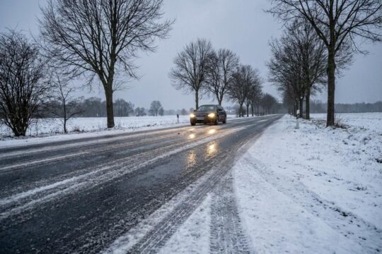 Vereiste, leicht schneebedeckte Landstraße in Niedersachsen, flankiert von Winterbäumen. Ein dunkles Auto fährt vorsichtig im trüben Winterwetter Niedersachsen.