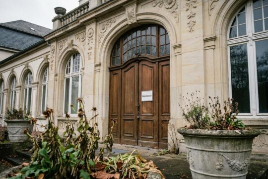 Die verriegelten Türen des eleganten Jugendstil-Eingangs symbolisieren die endgültige Reha-Zentrum Schließung Bad Nauheim. Welkende Pflanzen betonen die Melancholie.