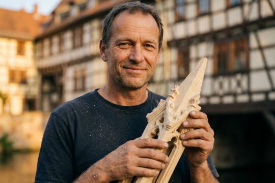 Auf der Krämerbrücke präsentiert Thüringer des Monats Martin Gobsch lächelnd eine kunstvolle Holzfigur, sichtbar gezeichnet von seiner Passion für das Handwerk.