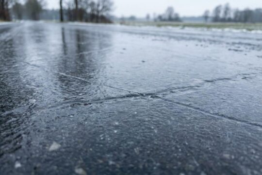 Dunkle Asphaltstraße, spiegelglatt, eisbedeckt mit Rissen. Glättegefahr in Niedersachsen. Unscharfe Winterlandschaft.