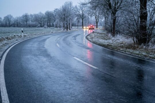 Winterliche Landstraße in Niedersachsen: Die dämmerige Fahrbahn glänzt tückisch, da unsichtbares Glatteis den Asphalt bedeckt. Karge Bäume mit Reif am Rand.