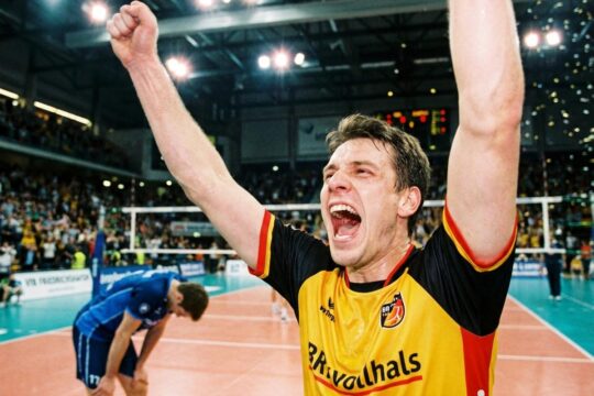 BR Volleys Spieler im Trikot jubelt ausgelassen mit erhobenen Armen über den BR Volleys Sieg gegen Friedrichshafen; ein frustrierter Gegner im Hintergrund.