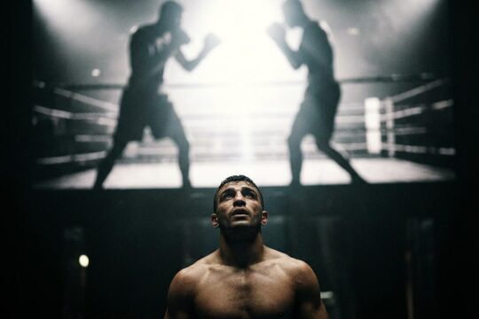 Ein muskulöser Boxer blickt entschlossen zu zwei schattenhaften Giganten auf, symbolisch für die Raymond Muratalla Herausforderung. Dramatisch beleuchtete Boxarena.