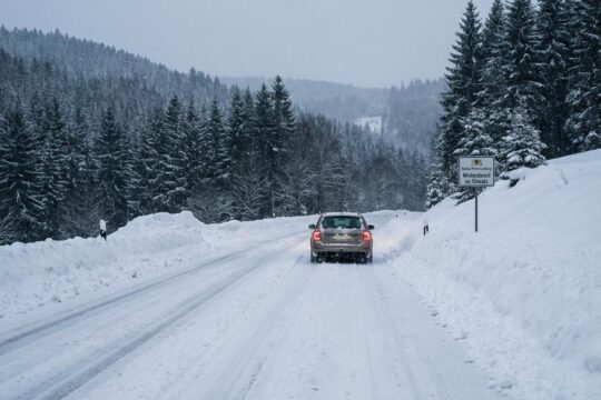 Tief verschneite Autobahn in Baden-Württemberg, umgeben von Tannenwäldern, wo sich ein einzelnes Fahrzeug durch den starken Schneefall kämpft.