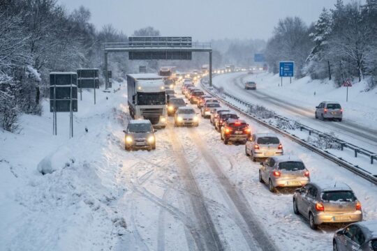 Dicker Neuschnee auf einer Autobahn in Hessen bei eisiger Morgendämmerung: Feststeckende Autos und LKW im Schneechaos, Lichter leuchten diffus.