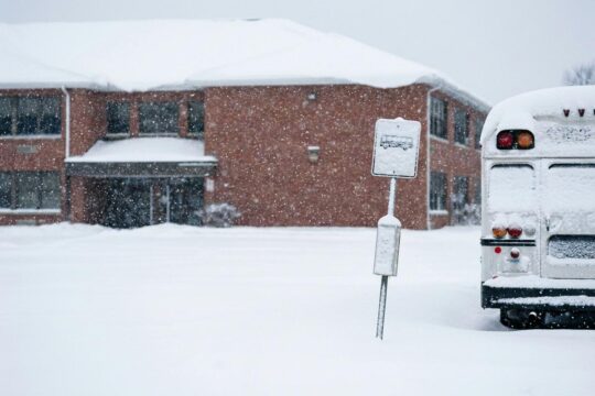 Ein menschenleerer, tief verschneiter Schulhof mit rotem Backsteingebäude und zugeschneitem Bushaltestellenschild veranschaulicht den Schulausfall Niedersachsen.