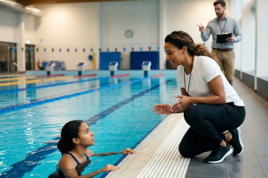 Ein Schwimmtrainer am Beckenrand gibt einem jungen Athleten Anweisungen. Im Hintergrund eine Person mit Tablet. Vielfältige Berufe im Schwimmsport.