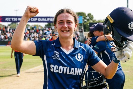 Junge schottische Cricket-Spielerin im blau-weißen Trikot feiert euphorisch den Erfolg der Schottland Cricket Frauen WM Qualifikation mit erhobenen Armen.