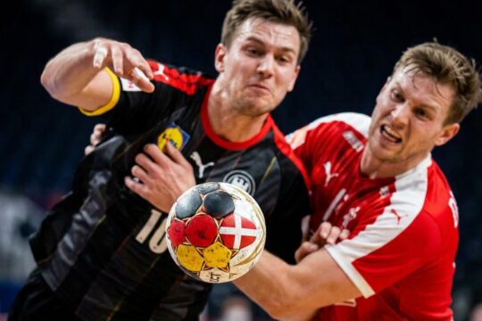 Ein Handball in Flugbahn, Spiegelungen deutscher und dänischer Farben, zwischen zwei fokussierten Spielern im spannenden Duell: Deutschland gegen Dänemark Handball.