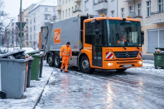 Ein BSR-Mitarbeiter steht auf eisiger Berliner Straße neben einem stillen Müllfahrzeug. Übervolle Tonnen zeigen, wie die Müllabfuhr bei Glatteis pausiert.