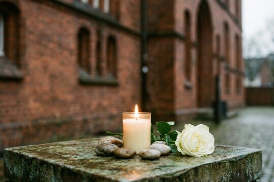 Feierliche Gedenkkerze, weiße Rose und Kiesel auf einem Stein vor historischem Backstein. Eine stille Erinnerung an den Holocaust-Gedenktag Schleswig-Holstein.