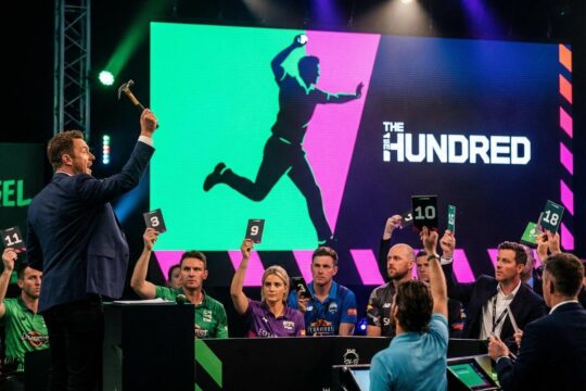 Ein Auktionator mit Hammer blickt auf konzentrierte Bieter bei der spannungsgeladenen The Hundred Spielerauktion. Im Hintergrund leuchtet eine Anzeigetafel.