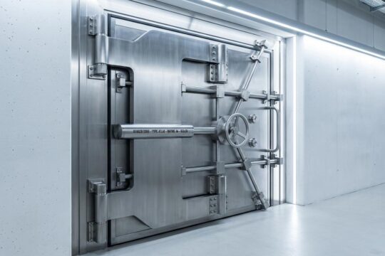 Moderne Schutzraumtür aus gebürstetem Edelstahl, massiv in Betonwand. Robuste Scharniere zeigen die hohen Anforderungen an Schutzraumtüren für Sicherheit.