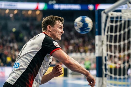 Ein konzentrierter deutscher Handballspieler wirft den Ball dynamisch aufs Tor, um den Einzug ins Handball EM Halbfinale Deutschland zu sichern. Die Arena ist erfüllt von Spannung.