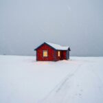 Eine tief verschneite Winterlandschaft mit einem warm leuchtenden Holzhaus, das die Essenz von Christophe Jacrot Winterland einfängt und Hoffnung im Eis spendet.