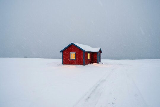 Eine tief verschneite Winterlandschaft mit einem warm leuchtenden Holzhaus, das die Essenz von Christophe Jacrot Winterland einfängt und Hoffnung im Eis spendet.