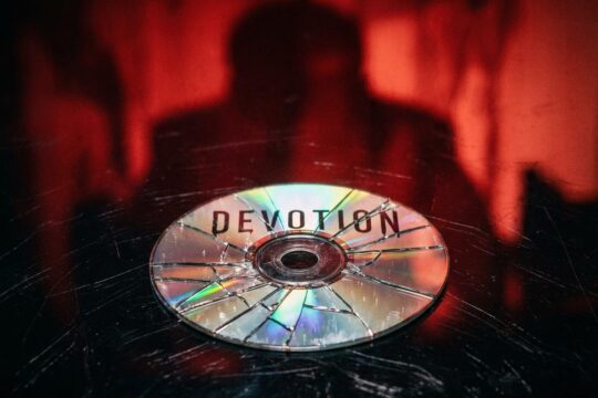GOG Devotion Kontroverse: Zersplitterte "Devotion" CD auf dunklem Grund symbolisiert Vertrauensbruch. Rote bedrohliche Silhouette im Hintergrund.