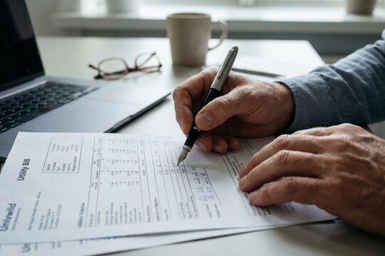 Hände prüfen akribisch eine detaillierte Nebenkostenabrechnung. Ein Stift markiert Posten auf dem Dokument auf einem aufgeräumten Schreibtisch im Büro.