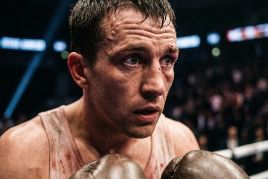 Josh Kelly unter Druck: Sein erschöpftes Gesicht, gezeichnet von Schweiß und Cut, zeigt angespannte Konzentration in der dunklen Boxarena.