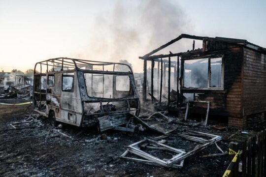 Das verheerende Nachspiel der Brandstiftung im Campingpark Geyer: Verkohlte Trümmer, Asche und aufsteigender Rauch in der Morgendämmerung, mit Polizeiband.
