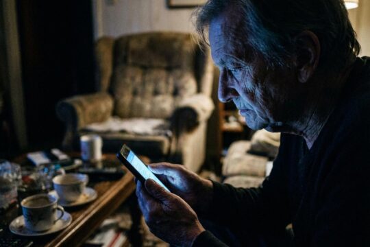 Eine ältere Person sitzt isoliert im dunklen Wohnzimmer, starrt auf das Smartphone. Das beleuchtete Gesicht zeigt die Symptome der Handysucht im Alter.