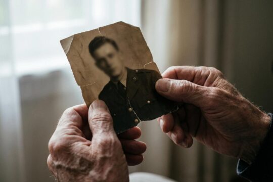 Nahaufnahme älterer Hände, die behutsam ein zerknittertes Sepiafoto einer undeutlichen Figur in dunkler Uniform halten. Der Sohn von SS-Mann reflektiert die Last seiner Herkunft.