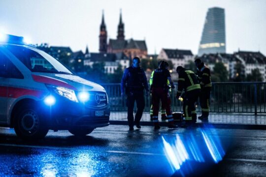 Nächtliche Szene in der Blaulicht Region Basel: Intensives Blaulicht spiegelt sich auf nasser Fahrbahn. Konzentrierte Einsatzkräfte agieren vor Basler Skyline.
