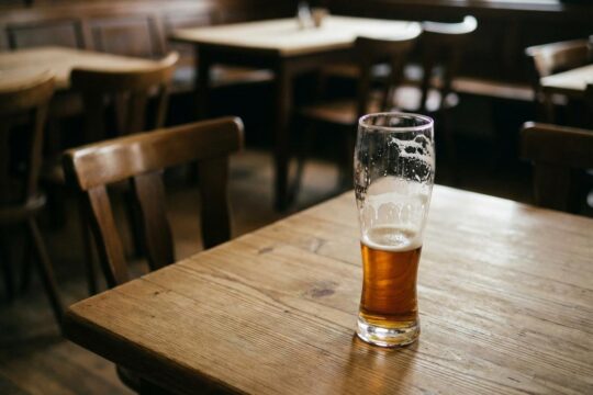 Ein halb volles Bierglas auf einem Holztisch in einer stillen Gaststätte, wo der Bierkonsum sinkt und Tische leer bleiben.