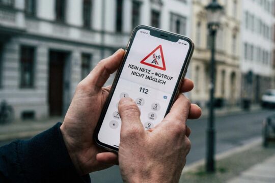 Verzweifelt drückt eine zitternde Hand auf ein Smartphone, das "112" und "Kein Netz" anzeigt, was ein Funkloch beim Notruf dramatisiert. Häuser im Hintergrund.