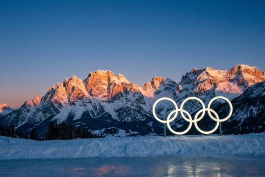 Helle Olympische Ringe ragen aus frischem Schnee vor den golden leuchtenden Dolomiten in Cortina d'Ampezzo. Ein Panorama zur Vorbereitung auf die Olympischen Winterspiele 2026.
