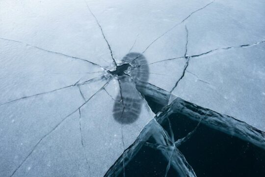 Nahaufnahme einer trügerisch glatten Eisfläche mit feinen Rissen, durch die dunkles Wasser schimmert. Ein Fußschatten zeigt die Gefahr beim Eisfläche betreten.