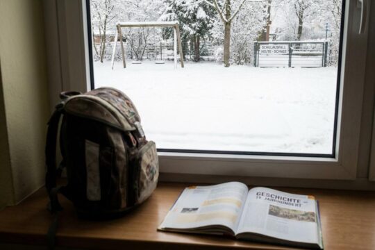 Schulranzen und offenes Lehrbuch liegen auf der Fensterbank. Draußen bedeckt tiefer Schnee die Straße und signalisiert Schulausfall wegen Schnee.