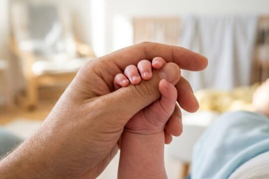 Eine schützende Erwachsenenhand hält sanft die winzige Babyhand, die den Zeigefinger umschließt. So erfahren junge Familien Hilfe durch Wellcome-Paten.