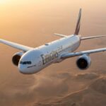 Eine gestreckte Boeing 777-10 in Emirates-Lackierung fliegt bei Sonnenuntergang über eine Wüste. Ihre Länge und die Triebwerke sind aus Untersicht betont.