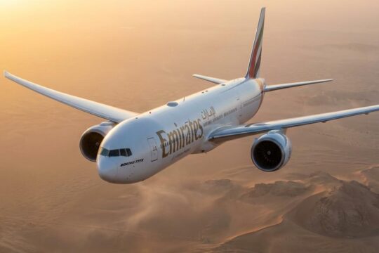 Eine gestreckte Boeing 777-10 in Emirates-Lackierung fliegt bei Sonnenuntergang über eine Wüste. Ihre Länge und die Triebwerke sind aus Untersicht betont.