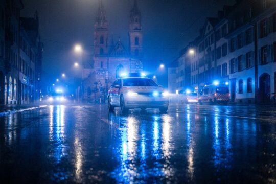 Dramatische Nachtszene in der Blaulicht Region Basel: Blaulichter eines Einsatzfahrzeugs spiegeln sich auf nassem Asphalt. Im Dunst weitere Wagen.